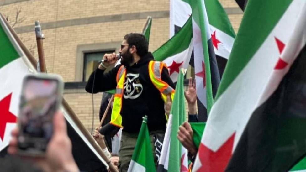 A man wears a reflective vest and holds a microphone as he's surrounded by people cheering, holding up Syrian flags and taking pictures.