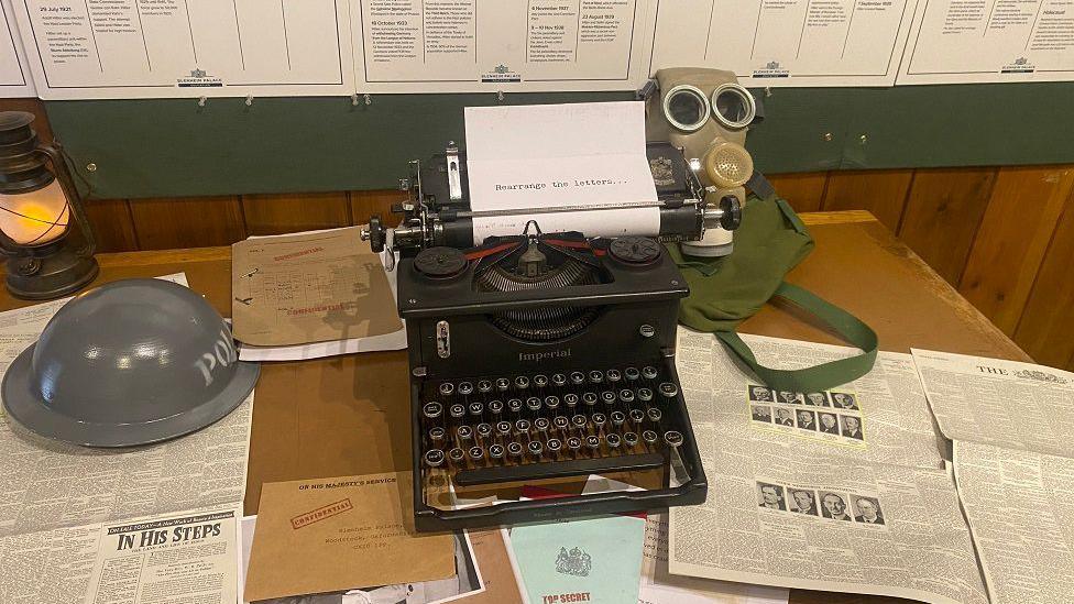 A photograph of part of the exhibition including newspapers spread out from the time, a typewriter, brown envelopes, metal grey hat, a gas mask and posters with writing on stuck on the wall.