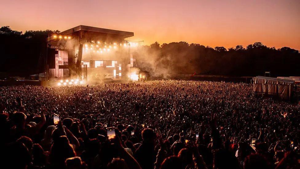 A teeming crowd of thousands holding up smartphones towards a stage lit up with yellow lights and covered with dry ice. A line of trees can be seen in darkness as the sun sets.