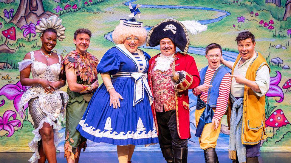 Simone Robinson as Wendy Darling/Myrtle, in a silver dress and headdress; David Breeds as Peter Pan in a leafy costume; Gordon Cooper as the dame in a curly blond wig, wearing bright make-up and a blue and white dress; John Thomson as Captain Hook, with a large black hat, red jacket and holding up his left arm with its hook hand; George Webster as Starky in a brown tricorne hat, red and white stripped top under a blue waistcoat, wearing clear plastic glasses and Oliver Scott as Smee, in a brown tricorne hat and yellow waistcoat over a white shirt.