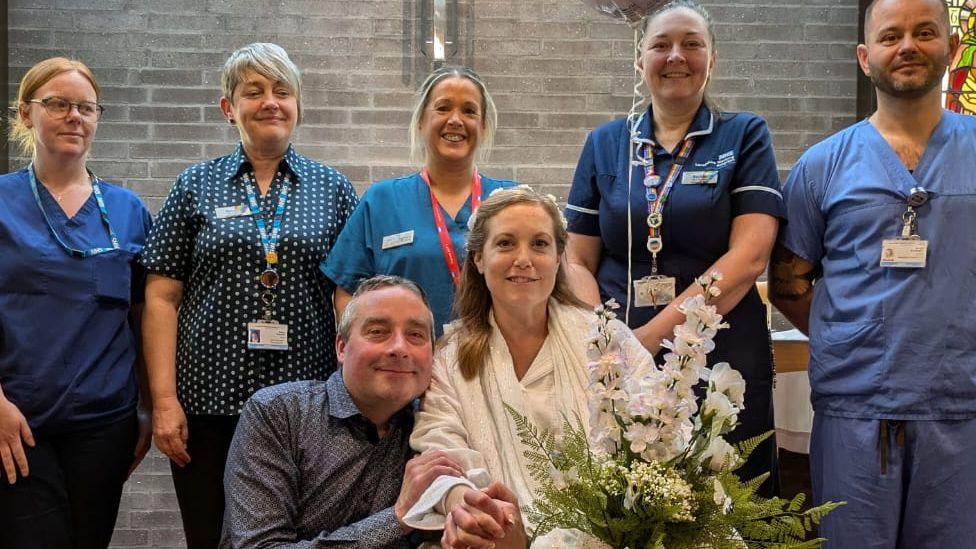 Emily and Jamie Cross  seated in front of four women and one man wearing blue hospital scrubs and nurses uniforms in the hospital chapel