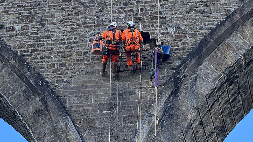 Repair work being carried out to a section of viaduct built of rough, blackened stone. Two workmen are on a suspended platform high up between two arches suspended by ropes from above, wearing orange trousers and coats and black harnesses. The sky to either side of the section of viaduct is cornflower blue.
