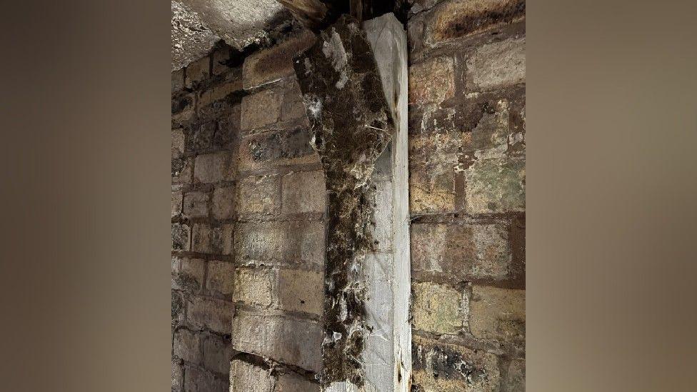 The black fabric remains of blackout curtains, that are nailed to a light wooden doorframe which is on a brick wall. The fabric is aged and in tatters, with white cobwebs all over it