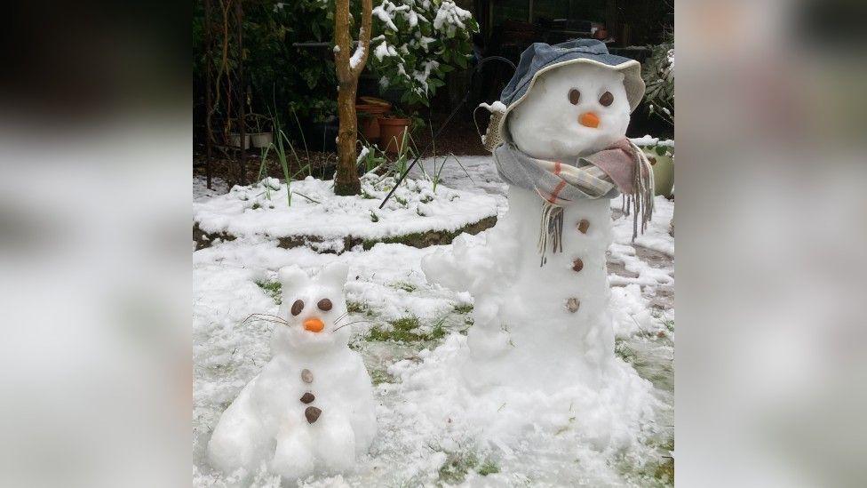 A snow figure shaped like a cat with rock eyes and stick whiskers, next to a snowman featuring a hat and scarf.