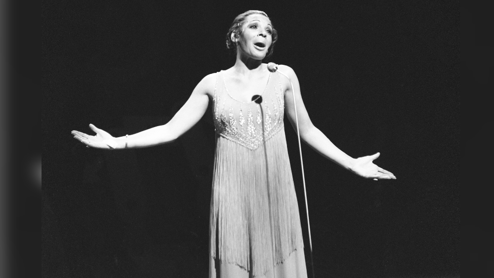 Shirley Bassey in a dress singing with a microphone in front of her