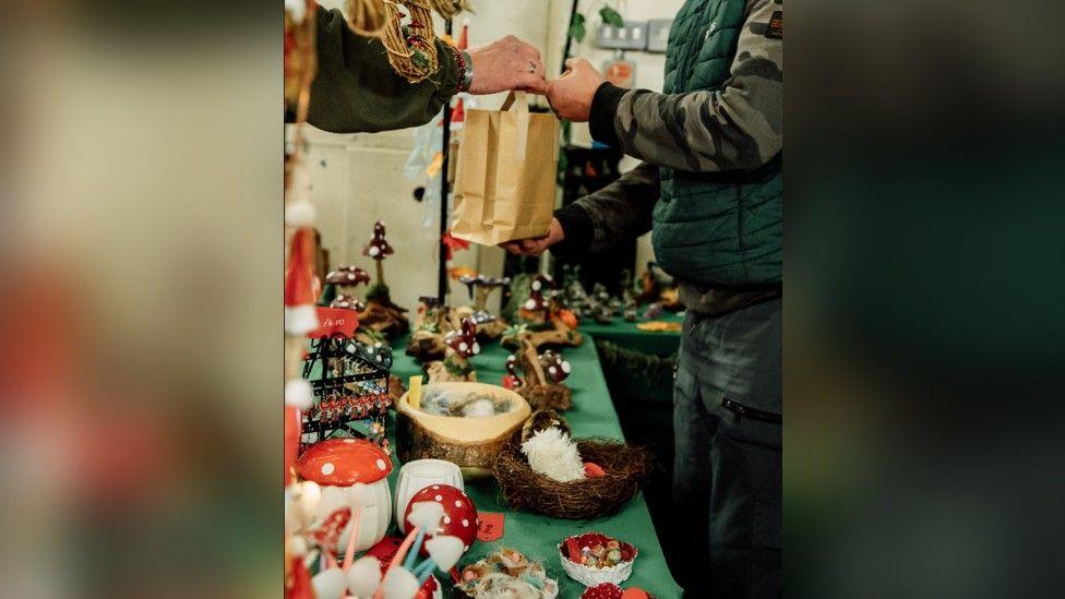 A market stall is selling mushroom themed items. A trader is handing over a brown paper bag to a customer.