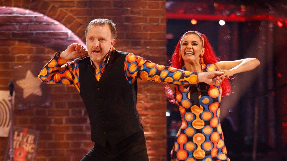 Chris McCausland in a bright shirt and black waistcoat costume, dancing with his partner on Strictly Come Dancing