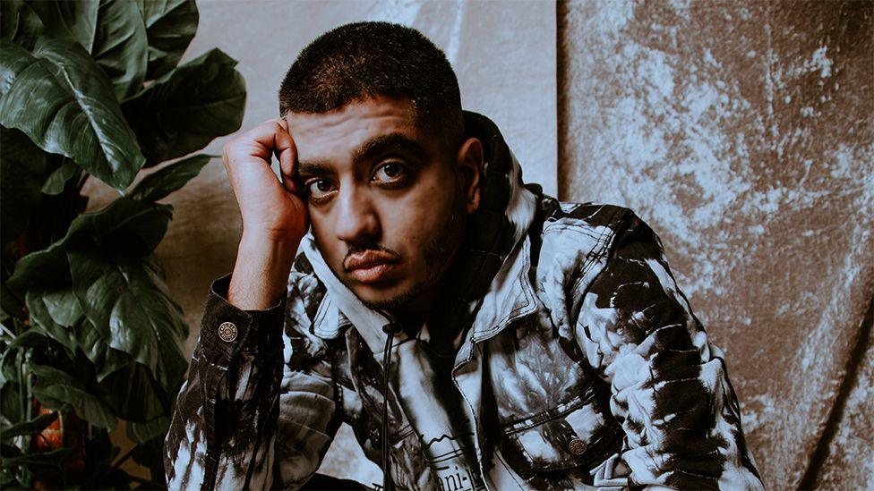 A man resting his head on his right hand, a black and white printed jacket. Behind him on the left is a green plant.