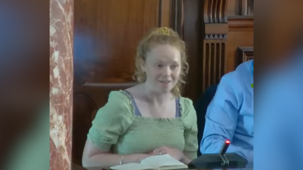The image shows a young woman with red hair and wearing a moss green top sitting in a wood panelled room with a notebook open in front of her. The arm of a man sitting next to her is visible.