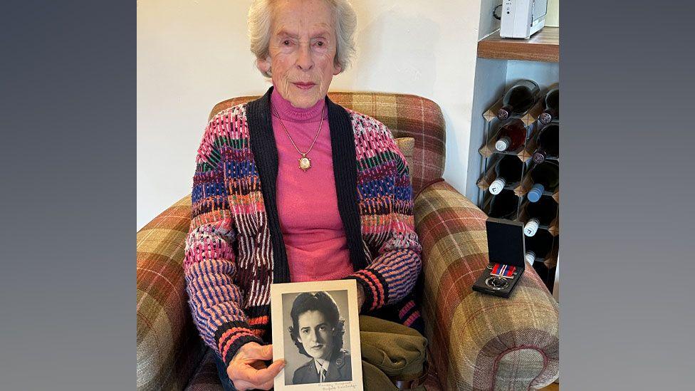 Joyce Wilding with grey short hair wearing a pink roll-neck under a multicoloured cardigan sitting on a plaid arm chair holding a World War Two era black and white photo of herself with a medal in its box on the arm of the chair