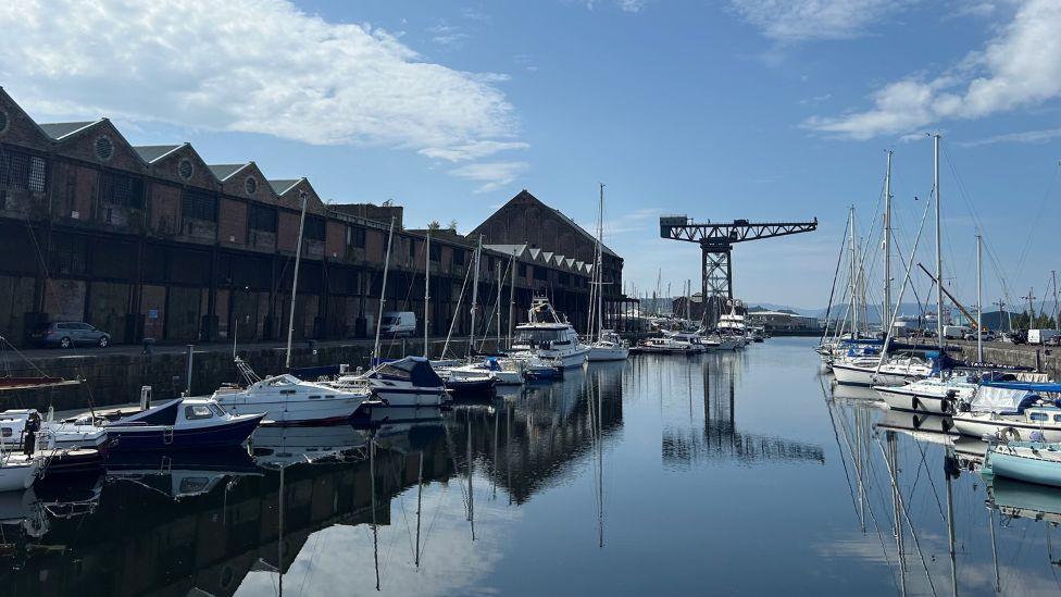 A marina with sailing yachts on a clear sunny day. At the end stands a large crane, with brick warehouse buildings on the left.