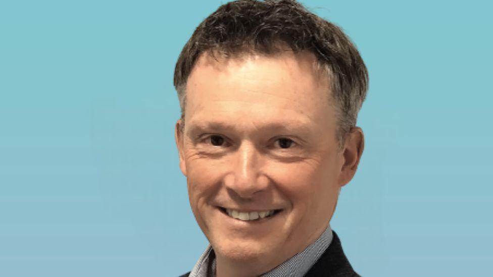 A man with dark hair smiles at the camera in a close-up shot in front of a blue background 