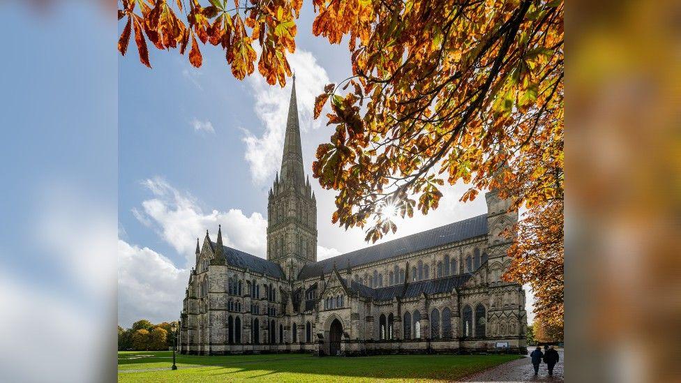 Salisbury Cathedral in the autumn sun, which is shining though red and brown leaves on a tree. There is bright blue sky, with a few wispy white clouds. 