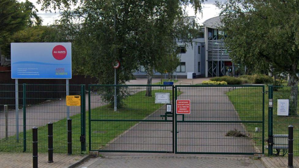 All Saints Academy's main entrance, showing a closed front metal gate across the road that leads to the main building.