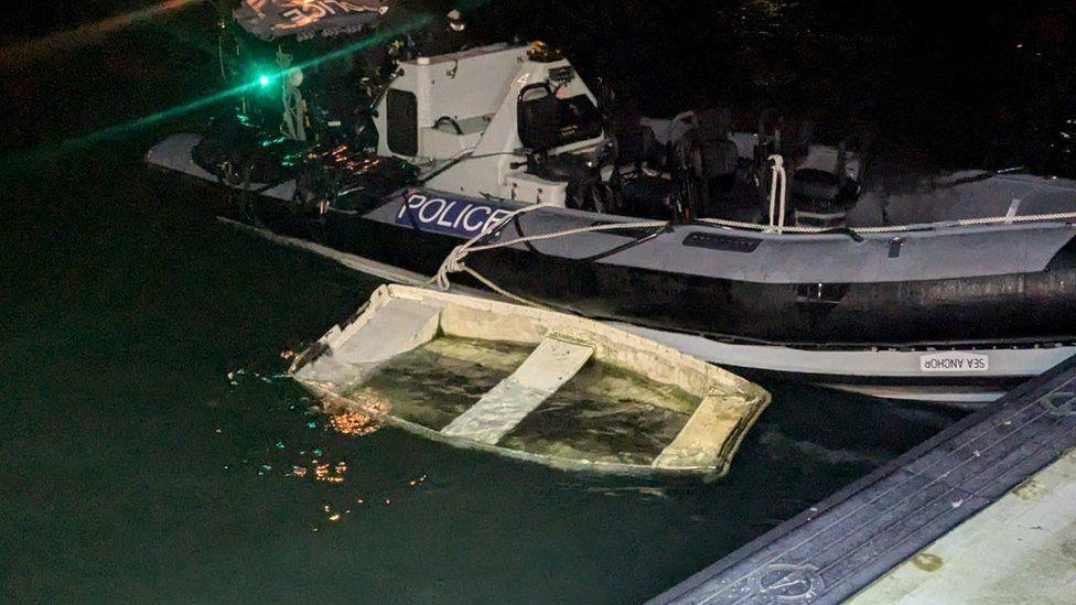 Grey and black rigid inflatable boat with green flashing light - small wooden white boat filled with water seen tied with rope to the police boat