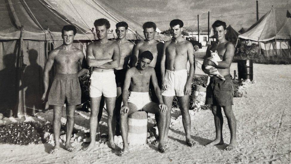 A black and white photo of seven young servicemen on Christmas Island in the 1950s. They are all wearing shorts and have no tops on. There are tents behind them.