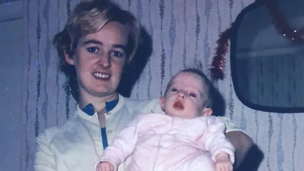 Bethan as a small baby being held by her mother in a living room.