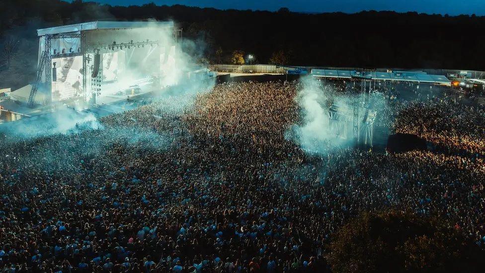 Mist wafts through the air above a crowds of thousands by a metal framed stage lit up at night in Heaton Park. 