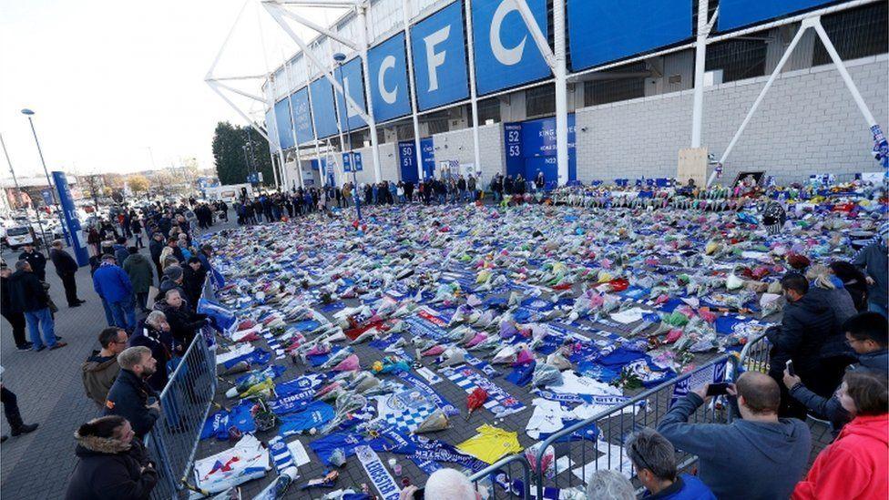 Rows of flowers and blue Leicester City shirts and flags laid out in front of a football stadium.