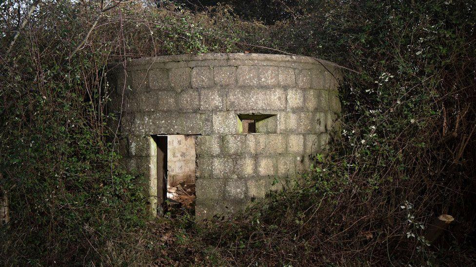 A round pillbox made from blocks of concrete. It has a rectangular door and a gun hole to the door's right. It is enveloped in brambles and ivy. 