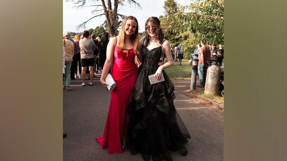 Ellie in a black dress smiling and looking at the camera stood next to her friend in a red dress. Both girls look very happy and are stood outside on their prom day.