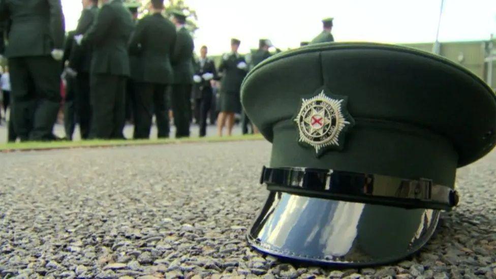 A PSNI hat sits on the ground n the foreground of the picture while male and female new police officers stand in the background in their uniforms