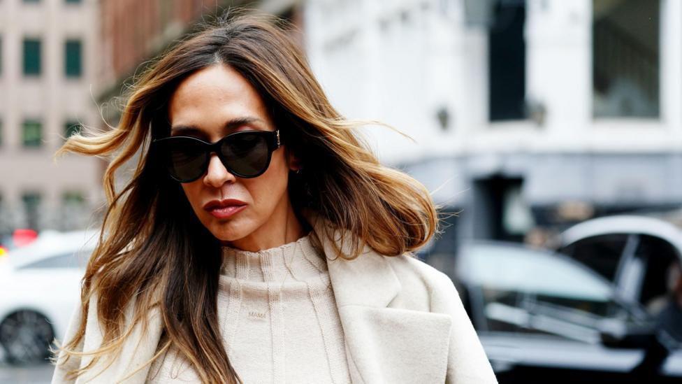 A woman with long brown hair is pictured wearing a cream-coloured coat and sunglasses.
