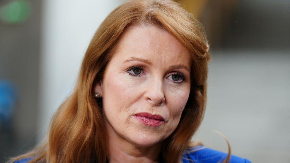 A woman with ginger hair, wearing a blue top, photographed in a close-up shot with a blurred background