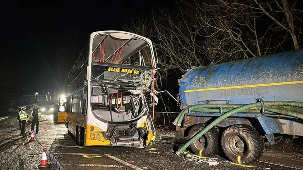 An image of the scene showing the damage to the school bus and the tractor, with the front of the bus smashed in. Police officers are present at the scene. 