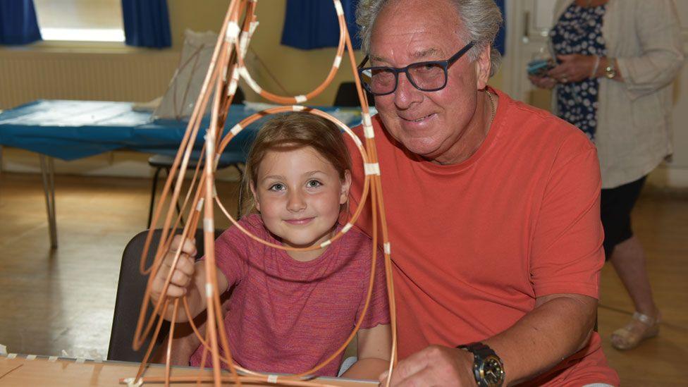 A grandfather in an orange t-shirt and granddaughter in a red t-shirt learning smiling as they learn how to make willow lanterns as in Eye, Peterborough