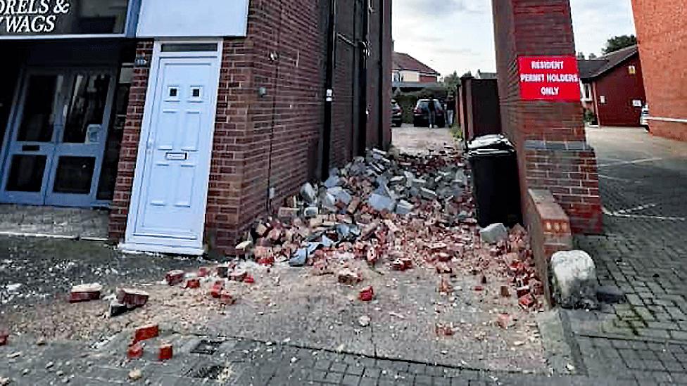 Bricks, rubble and black wheelie bins in a driveway. A white PVC door can be seen on a building to the left side of the driveway, and part of a shop sign to the door's left. To the right, a narrow red brick wall separates the driveway and a wider driveway. On the wall, a red rectangular sign reads in white letters RESIDENT PERMIT HOLDERS ONLY   