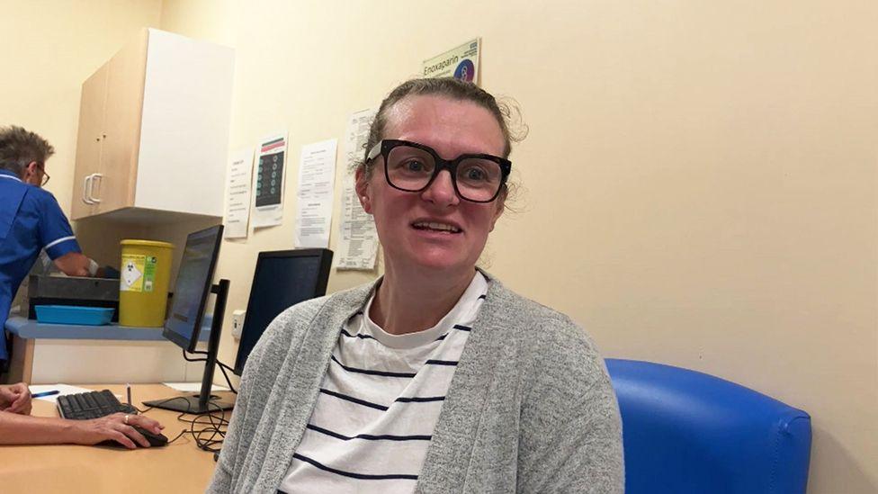 Pregnant Amy Louise Newell waiting for her vaccination she wears glasses and a white T-shirt with blue stripes and a grey cardigan