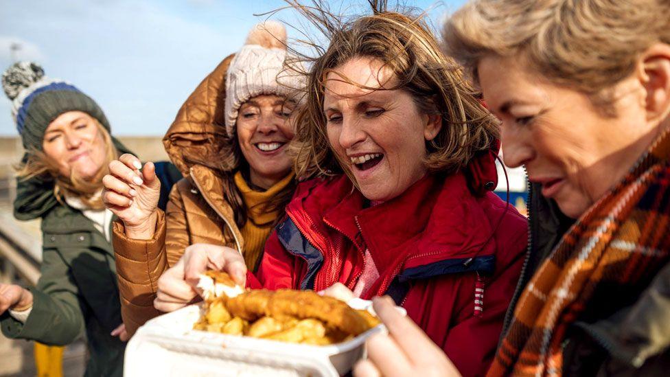 A group of four middle aged women wearing coats and hats as their hair blows in the wind tucking into a portion of fish and chips on the beach