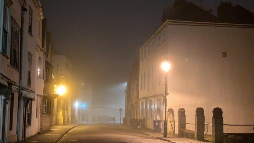 An empty street lined with houses  looking misty and illuminated only by two street lamps