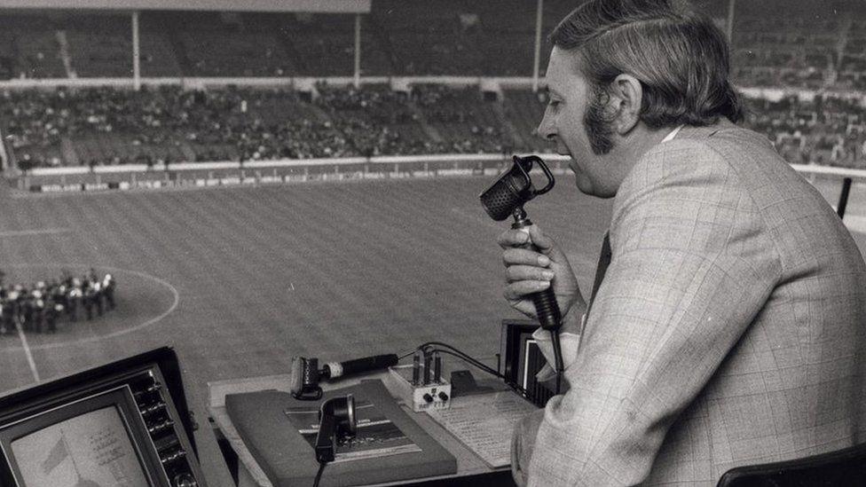 A black and white archived image of David Coleman wearing a checked patterned suit. He is speaking into an old microphone in a booth overlooking a football pitch. He has medium length hair which is pushed back behind his ears and thick sideburns. He is commentating on the match below.