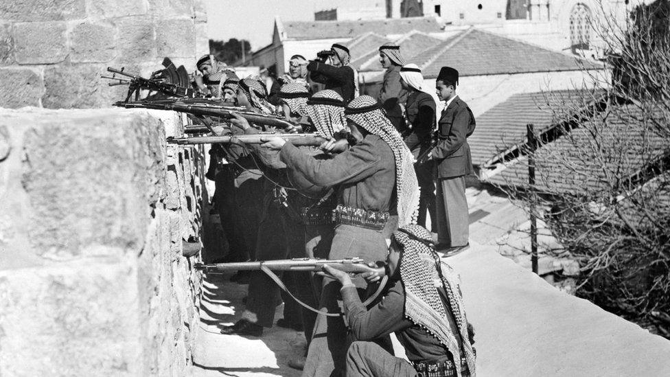 Arab Legion soldiers fire on fighters of the Haganah, the Jewish Agency self-defence force, in March 1948. A number of fighters are pointing rifles over a wall.