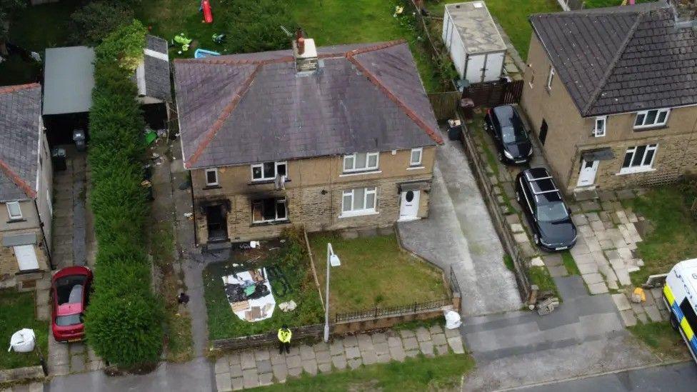 A burnt-out house, pictured from above. The front door and windows are covered with black fire damage and a police officer stands outside the property.