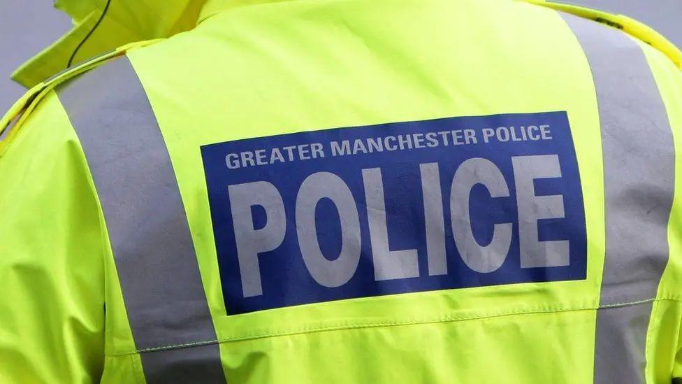 A Greater Manchester Police officer wearing a high-vis yellow jacket facing away in a zoomed in photograph.