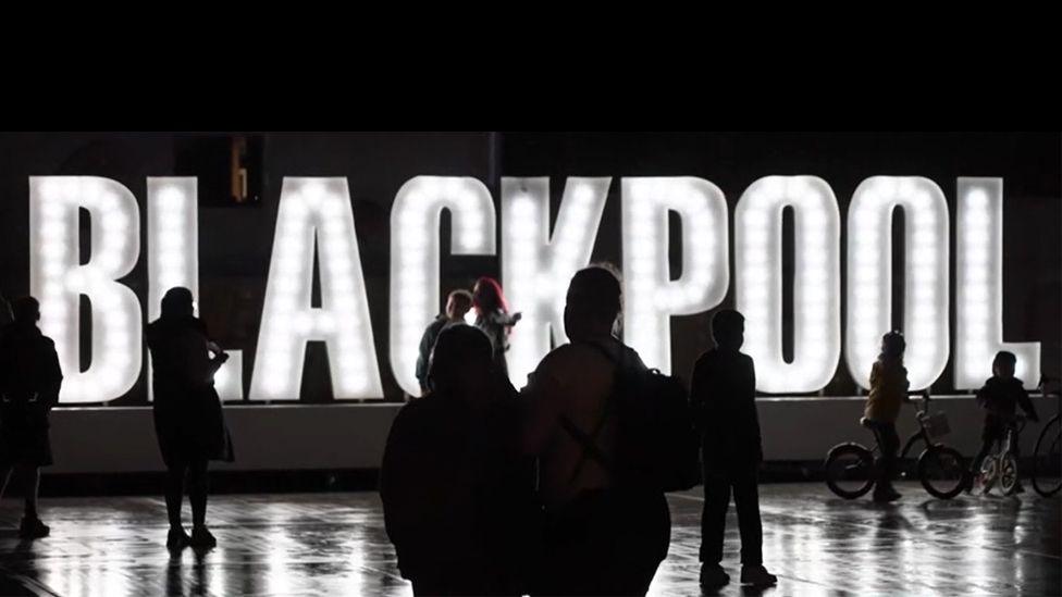 A large lit up sign with letters spelling out Blackpool in white while people in silhouette stand in front of it