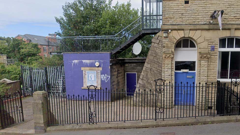 Street view of the outbuilding, a purple-painted square building at the side of the library surrounded by metal fencing