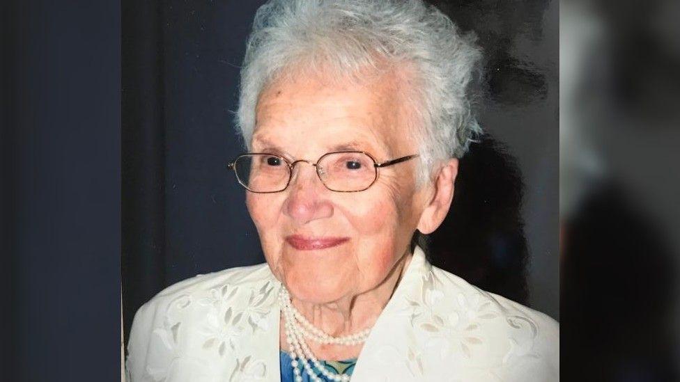 Mascha, pictured later in life. She is wearing a pale yellow jacket, pearls and a blue top. She also has metallic square glasses on, and has short grey hair. 