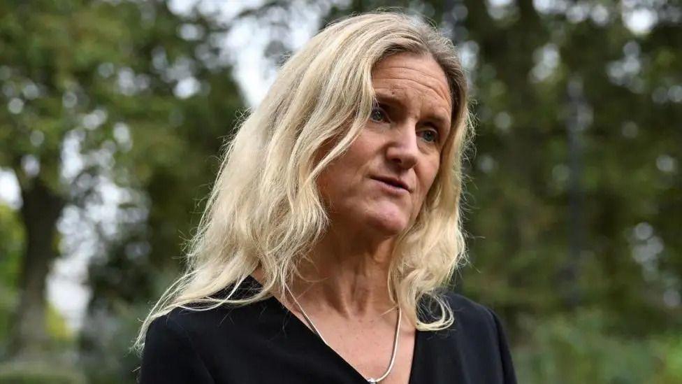 Kim Leadbeater looks to the left of the camera. She is wearing a black top and has a serious expression. She is wearing a gold necklace  The background is blurred, but very green, as she finds herself near a wooded area.