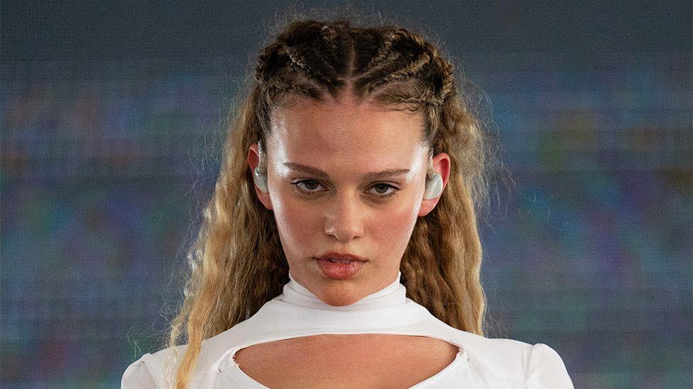 Elyanna performs during Austin City Limits Music Festival. She is wearing white, and has white ear pieces. The background is a screen showing ultraviolent.