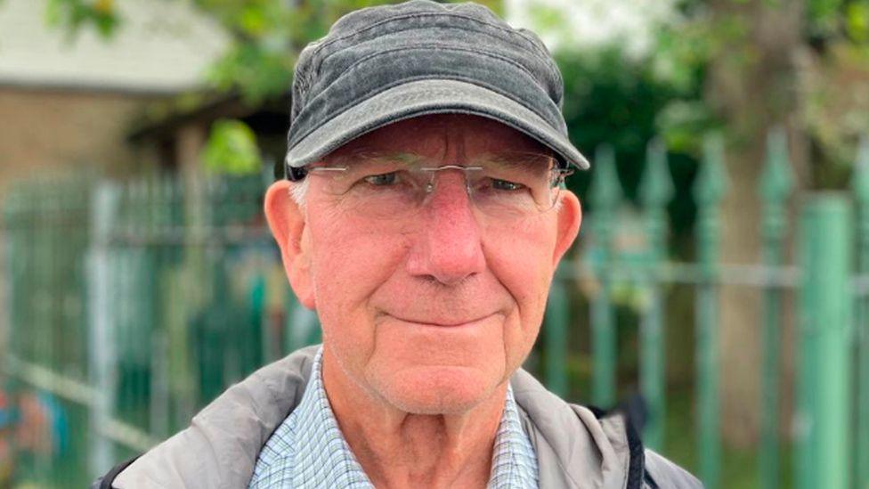 Head-and-shoulders shot of Richard Witts. He has short white hair under a faded grey baseball cap and frameless glasses. He is standing in front of green metal fencing with trees behind.