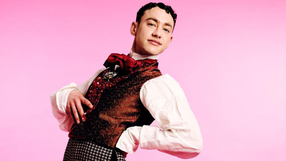Olly Alexander posing with his hand on his hip against a pink background in a promotional image for The Importance of Being Earnest