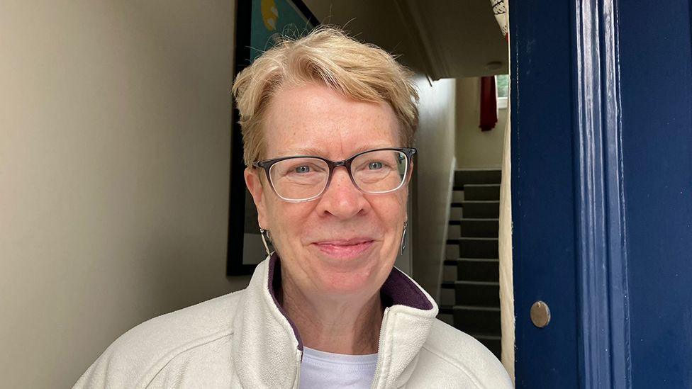Kate Sunley standing by the door of a house she has short blond hair and wears glasses and a white shirt and cream top
