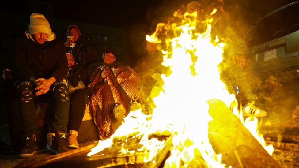 Migrants trying to reach the US gather in front of a bonfire to keep warm during a night of low temperatures in Ciudad Juarez, Mexico. They are wrapped up in coats and hats and huddle together. 