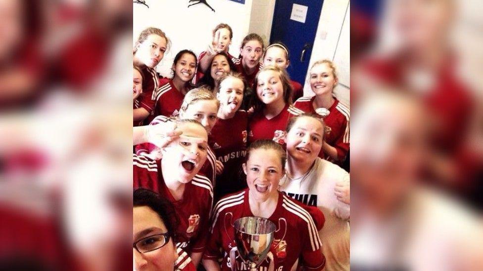 A joyous photo of a young women's football team in full kit in a changing room holding a trophy