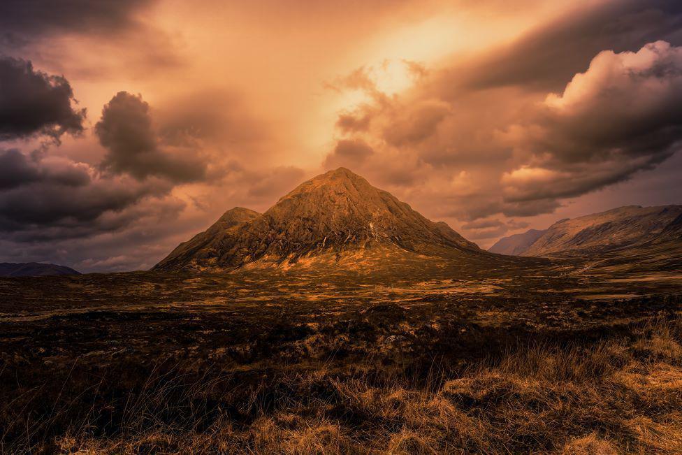 There is a bright light in the sky above the huge pointy mountain. There is grass in the foreground.