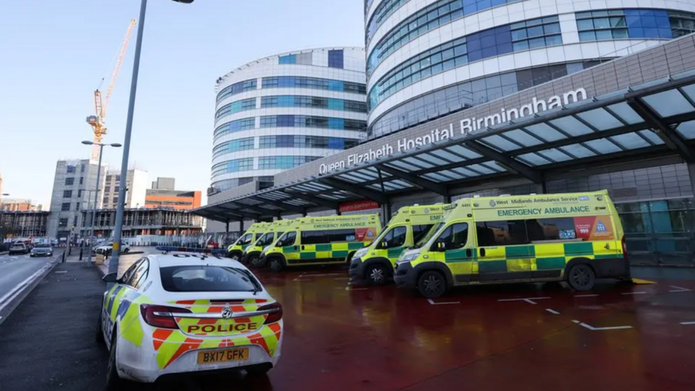 The front of a hospital building with a row of ambulances at the front and one police car. The sign above the door says Queen Elizabeth Hospital Birmingham.